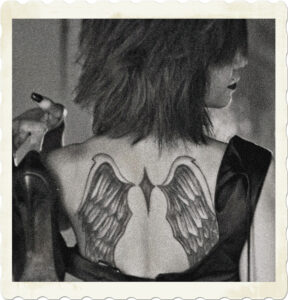 Picture of a woman with short black hair taken from the back. Tatoos of wings on her back is visible, and model is holding a pair of shoes. Image by Joey Velasquez from Pixabay.