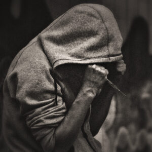 Picture of a man in a hoody, arms hiding his face. In his fist a syringe. Source Pixabay.com.
