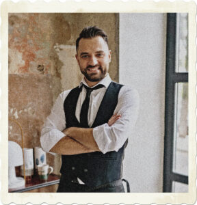 Image of a male server in a black and white uniform, with a trimmed beard, and smile. Image by Yan Krukov production from Pexels.