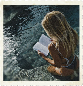Picture of a blonde sitting in shallow water of a pool. Facing away from the camera and wearing a bikini, they are reading a book. Image by Nastia Ligrain from Pexels.