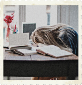 Picture of someone with long hair, slumped over a covered in open books as though taking a nap. Image by Andrea Piacquadio from Pexels.