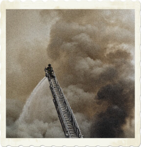 Picture of a firefighter atop a truck ladder spraying water below. In the background nothing by smoke is visible. Image by Brett Sayles from Pexels.