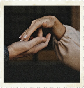 Picture of two hands touching. The one on the left is more masculine, no nail colour, and a formal jacket sleeve. One on right is feminine with a long sleeved blouse, wedding ring and coloured nails. Image by Nikolina from Pexels.