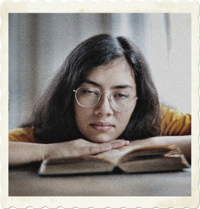 Picture of a woman with a book open. Her chin is resting on ehr hands, eyes half open, and neutral lips. Image by Andrea Piacquadio on Pexels.