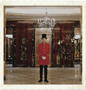 Picture of a man wearing a red long coat with a black tophat, standing in a rigid fashion in the centre of a hotel lobby. Image by Cottonbro Studio from Pexels.