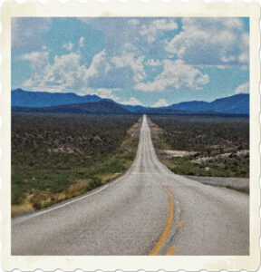 Picture of a two lane highway stretching out over plains towards mountains. There are no cars on the road, and the sky is cloudy. Image by MaxWdhs from Pixabay.