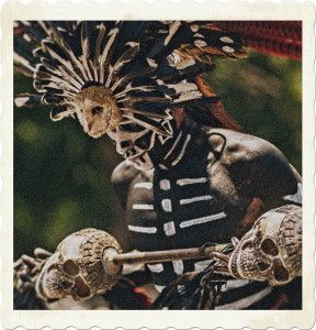Picture of a man painted to look like a skeleton, wearing ornate feathers, an owl head, and holding two carved skulls. Image by Alex Wolf (MX) on Pexels.