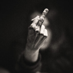 Picture focused on a hand with only the middle finger raised and a blurred woman in the background. Source Pixabay.com.