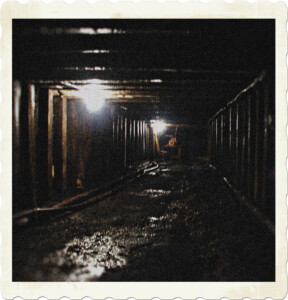 Picture of a coal mine shaft, featuring support beams, a dark and wet environment, and a few lights to shine the way.