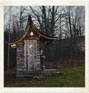 Picture of an outhouse in the woods, the wooden plans have faded from the sun, there's a pump on the side to wash, a moon cut into the door, and its lit from the inside.
