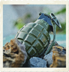 Picture of a pineapple-style grenade with spoon and pin in place amongst rubble. Image by D Alyansyah from Pixabay.