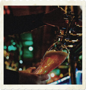 Picture of a bartender pouring a beer into a tall glass. The bar is dark with small light sources in the background. Picture by Darlene Alderson from Pexels.