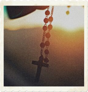 Picture of a rosary dangling from something, possibly a rearview mirror, with the sun in the background creating a silhouette of the item. Picture by Vanderlei Longo from Pexels.