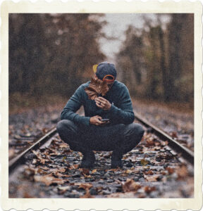 Picture of a squating on a railway line, holding a leaf to cover his face as he texts. Image by Craig Adderley from Pexels.