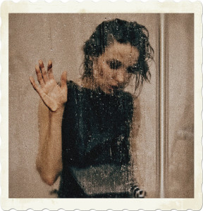 Picture of a woman in a shower, wearing a black dress and with wet hear. Her hand is pressing against the glass. Image by Marina Ryazantseva from Pexels.