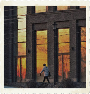 Picture of a woman in a puffy jacket walking in front of a building whose windows are relfecting a sunrise. Image by Screeny 42 from Pexels.