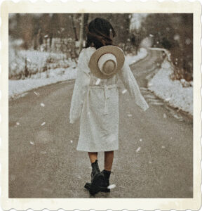 Picture of a brunette facing away from the camera. She is walking down a snowy windy road, wearing a long white coat, a cowboy hat hanging off her back and combat boots. Image by Jasmin Chew from Pexels.