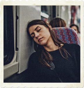 Picture of a woman sleeping against the bulkhead of a train. Other passengers are visible behind her, although blurry.