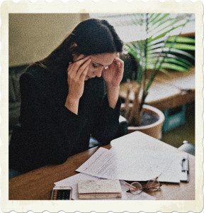 Picture of a woman at a desk covered in paperwork, rubbing her temples as though having a headache. Image by Leeloo Thefirst from Pexels.