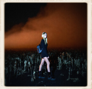 A young blonde woman with long straight hair, wearing white over-ear headphones, a black backpack, a short black dress, and shiny black patent leather platform ankle boots, stands in a field of dead, withered black plants under a thick rusty-orange fog at night.