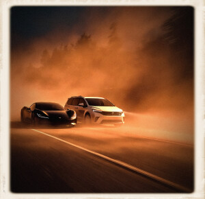 A sleek black near-future sports car and a white minivan race neck-and-neck at high speed on a remote highway at night, both cutting through thick rusty-orange fog with headlights glowing.