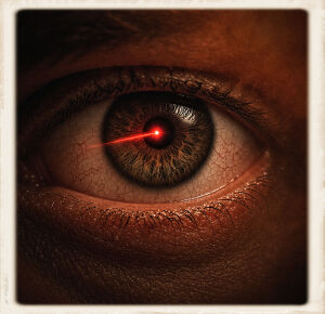 Extreme close-up of a human eye with a hazel-brown iris under reddish-orange lighting. A bright red laser beam ends in a sharp glowing point centered on the pupil. Detailed skin texture, fine wrinkles, and visible blood vessels in the sclera surround the eye. The frame has a soft polaroid-style border.