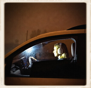 A young woman with blonde hair and white over-ear headphones drives a vehicle at night, viewed in profile through the side window. Soft interior light illuminates her focused expression as she holds the steering wheel, while thick rusty-orange fog surrounds the car outside.