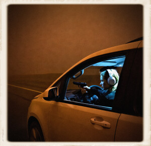 A young blonde woman wearing white over-ear headphones sits in the driver’s seat of a white vehicle at night, holding a black pistol aimed out the open side window while thick rusty-orange fog surrounds the car.