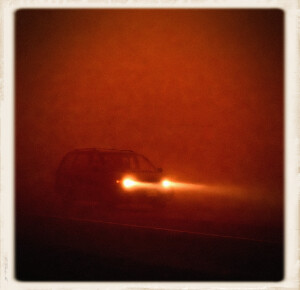A dark silhouette of a white minivan emerges from thick rusty-orange fog on a remote night-time highway, its headlights glowing brightly and casting beams into the dense haze.