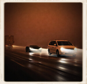 A white minivan leads a sleek black sports car on a foggy night highway, while a semi-tractor trailer passes in the opposing lane, all vehicles’ headlights cutting through thick rusty-orange fog.