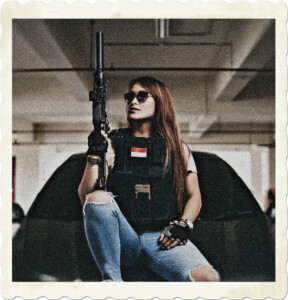 Picture of a brunette porting sunglasses, long hair and jeans. She is sitting on a black car in a parking garade wearing a kelvar vest and an assult rifle. Image by Nur Andi Ravsanjani Gusma from Pexels.