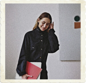 Picture of a dirty blonde wearing a long black collared shirt, touching her ear as though about to push back her heair. Eyes are mostly closed, smile looks natural, as though she is shy or embarassed. Holding portfolios and there appears to be a painting in the background. Image by Polina Zimmerman from Pexels. 