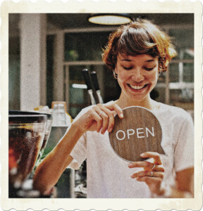 Picture of a short-haired brunette with a big smile and white shirt holding an open sign word bubble. There are coffe beans in the foreground and other coffee shop related material in the background. Image by Tim Douglas from Pexels.