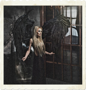 Picture of a blonde with long hair and a black dress fitted with wings that appear distorted, misshapen. Behind is a mirror, and the window to the side distorts the reflection.