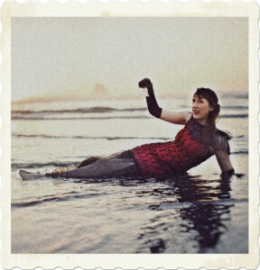 Picture of a flapper wearing a res dress, long black gloves, and holding a bell. Laying out in the water of a beach, during what appears to be a sunset. Image by Harmony Lawrence from Pixabay.