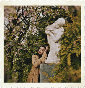 Picture of a brunette wearing a long coat, with eyes closed and red lips leaning against the statue of an angel. She is surrounded by trees, including flowering trees. Image by Maksim Goncharenok from Pexels.