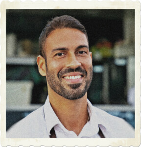 A potrait of a bearded man. Subburned skin, dark hair, neatly trimmed bears and white teeth. A blurry background of a restaurant or food truck in the background. Image by Kampus Production from Pexels.