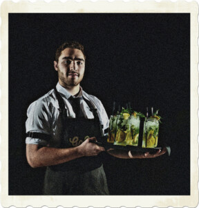 Picture of a bearded server wearing a white shirt, tie, black apron, and holding a bunch of flashy drinks. Image by Julio Irrazabal from Pexels.