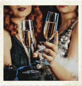 Picture of what appears to be two flappers whose faces are blurred in the background with their glasses of champage in focus.