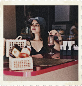 Picture of a woman sitting of a counter of a restaurant. Wears a baseball cap, long flowing brown hair, and a short top. Holds the straw to her drink, while looking out towards the entrance. Image by Alexandre Mattos from Pexels.