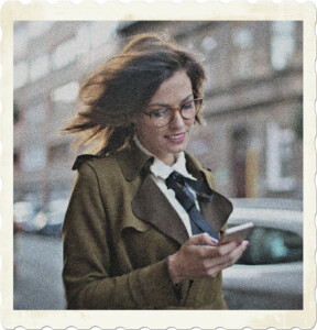 Picture of a brunette with glasses walkign down the streets. Wind is blowing their her hair, and she's smiling while looking at something on her phone. Image by Andrea Piacquadio from Pexels.