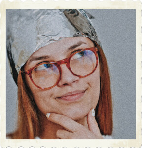 Picture of a redhead with red round glasses, looking up at the ceiling while pondering. There is a smirk on her face and a tinfoil hat to complete the ensemble. Image by Dids from Pexels.