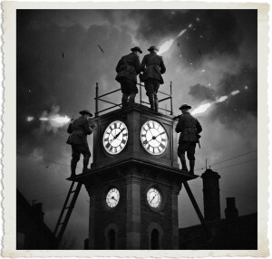 Four soldiers in Brodie helmets defend a tall brick clock tower at night during an intense battle. Two stand atop the platform while others position on the sides, firing weapons amid a dramatic sky filled with smoke, tracer fire, and falling debris. The black-and-white image features a vintage distressed photo border.