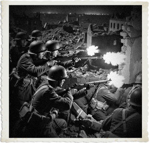 section of Canadian Expeditionary Force soldiers in 1915 khaki uniforms and Brodie helmets fire their rifles from behind rubble in a bombed-out town at night. Bright muzzle flashes light their faces and the surrounding debris. Black-and-white period photograph.