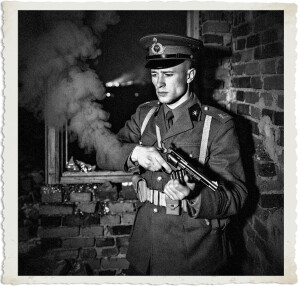 Canadian Expeditionary Force Captain Marc stands with his back against a brick wall at night, calmly reloading a Webley revolver. Thick smoke pours from the shattered window beside him. He wears a 1915 CEF officer’s peaked cap and khaki tunic with captain’s rank stars. Black-and-white period photograph.