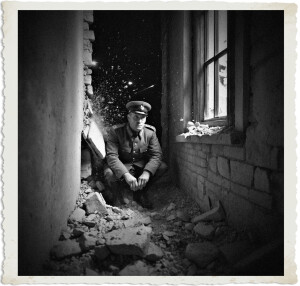 An officer in a peaked cap crouches tensely amid rubble inside a shattered building, as a violent explosion sends bricks and debris flying through the air around him. The stark black-and-white image captures the raw shock of battle, framed with a vintage distressed photo border.