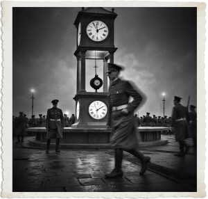 Black and white photograph of a large, ornate clock tower at night, surrounded by soldiers in military uniforms. A soldier in the foreground is captured in motion blur as he walks briskly past the tower, creating a sense of urgency. Other soldiers stand stationary in the midground and background near the base of the tower. The wet cobblestone ground reflects the lights, adding to the dramatic atmosphere. The image has a vintage, torn-edge photo border.
