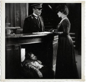 A dramatic black-and-white vintage-style photograph with torn paper edges, depicting a tense 1915 night scene in an elegant hotel lobby. A uniformed night concierge stands behind the polished reception desk, calmly conversing with an elegantly dressed socialite in a long dark gown, hat, and gloves. A dead man’s body is crudely crammed and partially visible in the cramped space beneath the counter. The image has heavy film grain, deep shadows, and high contrast, enhancing the unsettling, matter-of-fact horror.