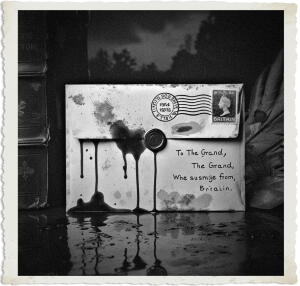 A dramatic black-and-white vintage-style photograph with torn paper edges, showing a close-up of a weathered 1915 British envelope resting on a dark, reflective surface. The envelope features a King George V postage stamp, a circular postmark, and a wax seal, with thick black ink (or blood-like liquid) dramatically dripping down from it and pooling below. Handwritten text on the envelope reads “To The Grand, The Grand, Whe susmje from, Britain.” The moody, high-contrast composition with deep shadows evokes supernatural dread.