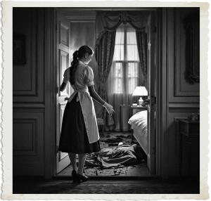 Black and white photograph of a maid in a classic uniform standing in a bedroom doorway, her back to the viewer. She looks into the room where a body lies face-down on the floor in a large pool of blood near an unmade bed. The vintage-style image has a distressed, aged border.